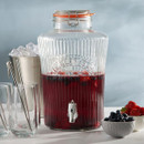 Large Kilner glass drinks dispenser filled with red berry punch, shown with glasses, ice bucket, and bowls of strawberries and blueberries.