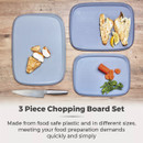 Three grey chopping boards on a wooden surface, each with food: cooked chicken, fish with lemon, and sliced carrots with broccoli. A knife is nearby. Text reads "3 Piece Chopping Board Set.