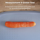 A bright orange carrot lies horizontally on a blue grater tool with embossed measurements. The wooden table background adds a rustic touch.