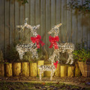 Illuminated outdoor reindeer family Christmas decorations with warm white LED lights and red bows, displayed in a garden against a wooden fence.