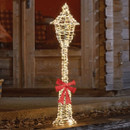 Illuminated outdoor Christmas lamppost decoration with warm white LED lights and a red bow, placed beside a wooden cabin door.
