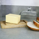 A block of butter resting on a wooden base with the ribbed glass butter dish lid removed and placed behind, next to slices of crusty bread on a grey kitchen worktop.