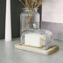 A rectangular glass butter dish with ribbed detailing and a wooden base, holding a block of butter. A tall glass vase with dried flowers is in the background.