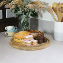 A wooden cake stand topped with a Victoria sponge, chocolate brownies, and iced sponge slices, styled without the dome cover on a kitchen worktop.