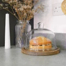 A round glass cake dome with wooden base, covering a Victoria sponge cake on a grey worktop with kitchen accessories.