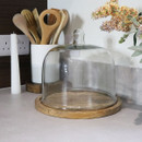 An empty large round glass cake dome with wooden base, placed on a kitchen counter alongside utensils and decorative accessories.