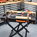 A workbench in a workshop with tools like a saw, screwdriver, hammer, and clamps on it. Metal drawers and wooden surfaces are in the background.