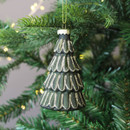A dark green Christmas tree-shaped ornament with gold glitter detailing, hanging from a green fir branch adorned with soft, glowing fairy lights.