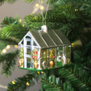 A silver greenhouse-shaped Christmas tree ornament hanging from a green fir branch. The decoration features gold flowerpots, green plants with red glitter details, and a silver roof, with soft fairy lights in the background.