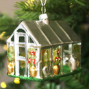 A close-up of a silver glass greenhouse Christmas ornament decorated with gold pots, green plants, and red glitter flowers, hanging from a Christmas tree branch surrounded by festive lights.