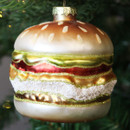 A close-up of the glittering burger-shaped Christmas ornament, showing its detailed layers of bun, lettuce, tomato, cheese, and patty, against the green pine needles of the tree.