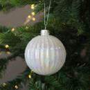 A glass iridescent Christmas bauble with a ridged design, hanging from a green Christmas tree branch with warm fairy lights in the background.