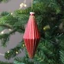 A red glass Christmas decoration with a ridged, elongated teardrop shape, detailed with gold glitter. It hangs from a green Christmas tree branch with warm fairy lights in the background.