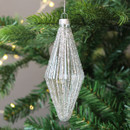 A silver glass Christmas tree ornament, covered in fine glitter and hanging from a green fir branch with softly glowing fairy lights in the background.