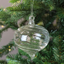 A clear glass Christmas bauble with a swirled onion shape, hanging from a silver thread on a green fir branch with soft fairy lights in the background.