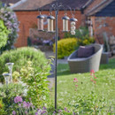 Four bird feeders hang from a metal pole, surrounded by a garden with colourful flowers. There is a wicker chair and a brick house in the background.
