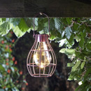 Hanging lantern with glowing bulb in a rustic cage design, suspended from a wooden beam amid lush green leaves, creating a warm, serene ambiance.