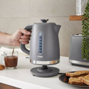 A hand lifts a sleek, grey electric kettle from its base on a kitchen counter. Nearby are brown toast slices and a half-full cup of coffee, creating a cosy morning scene.