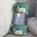 Rolled flannel blanket with a teal Highland cow print, wrapped in a grey Country Club label, displayed on a cosy sofa.