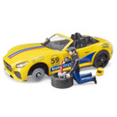Toy scene with a yellow convertible car and a figure kneeling beside it, changing a tire. The car features racing decals and a helmet nearby.