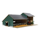 A model farm building with a green exterior has an open side showing cows inside. A red tractor with hay bales is parked under a canopy. The scene is tranquil.