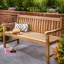 A teak outdoor bench with curved slatted back and wide armrests, styled with a cream cushion, positioned on a patio surrounded by lush garden foliage.