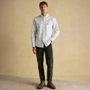 Man wearing a white Joules checkered shirt and dark green pants, standing against a light beige textured wall. The tone is casual and stylish.