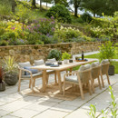 A modern wooden outdoor dining set arranged on a stone patio, featuring a large rectangular table and cushioned rope-woven chairs, surrounded by colourful borders and a rustic stone wall.