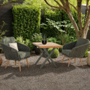 Bistro set in a garden with two dark green rope-woven armchairs and a square wooden-top table on crossed metal legs, surrounded by lush greenery and gravel flooring.
