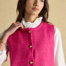 Close-up of a bright pink knitted vest showing the rounded neckline and decorative gold buttons over a white ruffled blouse.