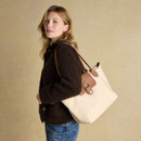 Cream tote bag with long tan shoulder straps and a front tan flap detail, worn over the shoulder by a woman in a dark brown textured jacket and blue jeans.