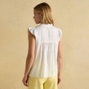 Back view of a woman wearing a sleeveless white blouse with ruffled cap sleeves and a softly gathered yoke, styled with yellow embroidered shorts against a beige backdrop.