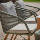 Detail of a rope-woven outdoor dining chair with a pale cushion and wood effect legs, showing the textured armrest and backrest.