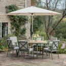 A round wood effect garden dining table with six sage-green cushioned chairs beneath a cream parasol on a stone patio beside a cottage wall.