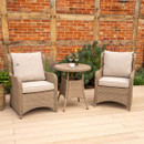 Outdoor beige rattan bistro furniture with two cushioned armchairs and a round wicker table, shown on decking with a rustic brick wall backdrop.