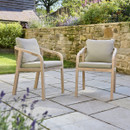 Pair of wooden outdoor dining chairs with rope detailing and grey cushions on a stone patio in a garden setting.