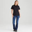 Full-length view of a woman wearing a fitted black polo shirt styled with blue jeans and dark ankle boots. The shirt features a classic collar, button placket, and subtle chest logo detail.