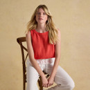 Woman seated on a wooden chair wearing a sleeveless red linen top with white drawstring trousers, styled in a relaxed summer look.