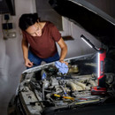 A woman examines a car engine with a bright, adjustable work light illuminating the area. She's holding a blue cloth, conveying focus and determination.