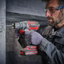 A person in safety glasses and gloves uses a red cordless drill on a concrete wall. The scene conveys focus and precision in a construction setting.