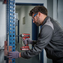 Man wearing protective gear uses a red power drill on a blue metal shelving unit in a workshop. He appears focused and precise in his work.