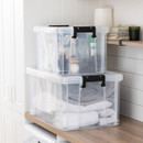 Stacked transparent storage boxes with clip-lock handles holding household cleaning items and folded towels in a utility room.