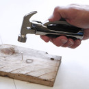A hand holds a versatile multi-tool hammer above a wooden plank with a nail. The setting suggests a DIY project, conveying focus and practicality.