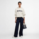 Full-length view of model in an off-white Barbour logo hoodie with wide-leg dark indigo jeans, carrying a denim tote bag.