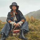Model seated outdoors in grassy hills wearing a dark waxed jacket, wellington boots and a bucket hat with chin cord, holding a crossbody bag.