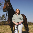 Outdoor lifestyle shot of a woman in a sage green Barbour half-zip sweatshirt standing beside a horse in a field.