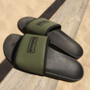Olive green Barbour slider sandals with wide branded strap and black contoured footbed, photographed on a wooden railing outdoors.