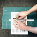 Hands holding a cutter and ruler, trimming a barcode paper on a green cutting mat. The setting conveys precision and focus in a crafting task.