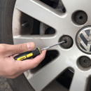 A person uses a small screwdriver to remove a wheel nut cover from a car tire. The image shows a close-up of the hand and the alloy wheel.