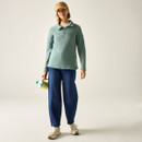 Full-length view of a model wearing an ivy green half-zip fleece top with blue jeans and cream trainers, holding a bottle.